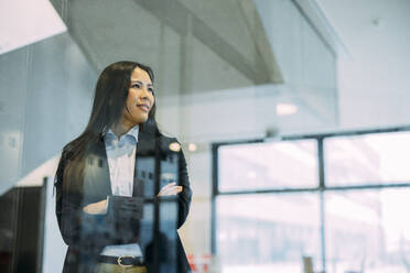 Businesswoman with arms crossed seen through glass at workplace - JOSEF21419