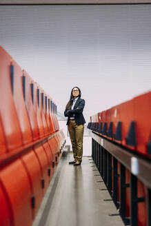 Confident businesswoman with arms crossed standing amidst red seats at convention center - JOSEF21446