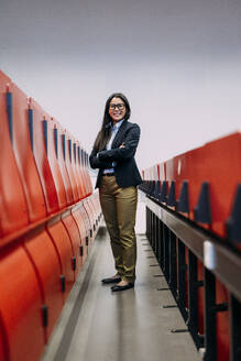 Smiling businesswoman with arms crossed standing amidst red seats at convention center - JOSEF21447