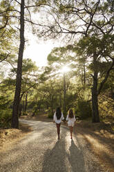 Teenage girl holding hands with mother walking on road in forest at sunset - ANNF00478