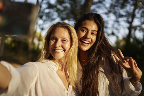 Happy woman taking selfie with teenage daughter showing peace sign in forest - ANNF00518