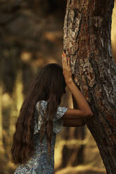 Teenage girl with long black hair leaning on tree trunk in forest at sunset - ANNF00535