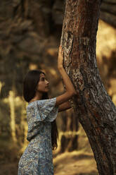 Teenage girl leaning on tree trunk in forest at sunset - ANNF00536