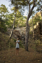 Teenage girl standing near trees in forest at sunset - ANNF00538