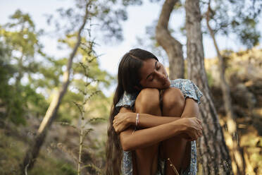 Lonely teenage girl with eyes closed hugging knees in forest - ANNF00542