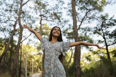 Smiling teenage girl with arms outstretched in forest at sunset - ANNF00553