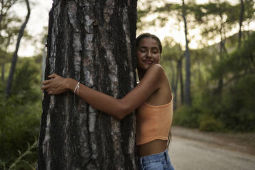 Smiling teenage girl hugging tree trunk in forest - ANNF00563