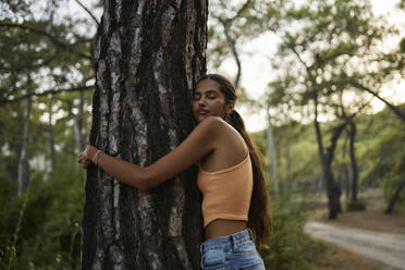 Teenage girl with eyes closed hugging tree trunk in forest at sunset - ANNF00564