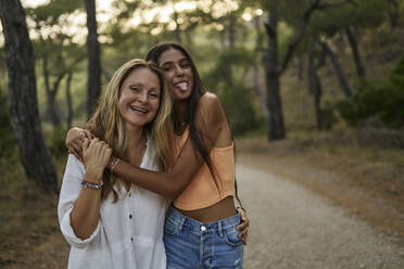 Happy teenage girl sticking tongue out hugging mother in forest - ANNF00573