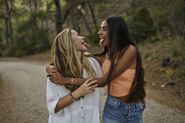 Happy mother and daughter looking at each other and sticking tongues out in forest - ANNF00577