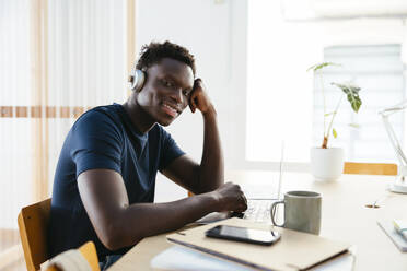 Lächelnder junger Student mit drahtlosen Kopfhörern und Laptop am Schreibtisch sitzend - EBSF03879