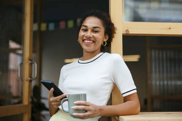 Lächelnder Student mit Kaffeetasse und Smartphone in der Tür stehend - EBSF03910