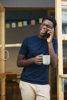 Glückliche junge Studentin steht mit Kaffeetasse und spricht auf Smartphone - EBSF03913