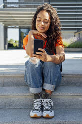 Smiling student using smart phone sitting on steps in campus - PGF01680