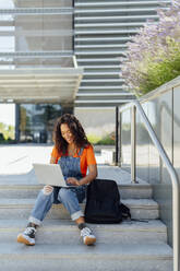 Happy student using laptop sitting on steps in campus - PGF01683