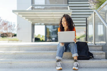 Smiling student sitting with laptop on steps in campus - PGF01684