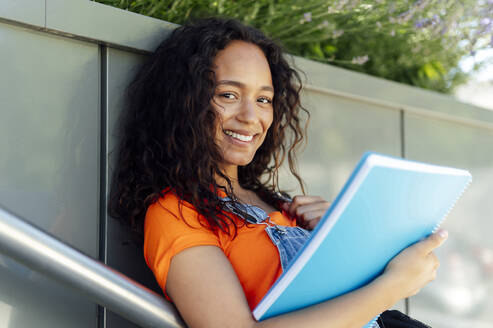 Smiling student holding spiral notebook near wall in campus - PGF01686