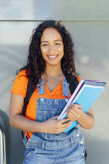 Happy student holding spiral notebooks in front of wall at campus - PGF01689