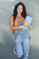 Smiling student holding spiral notebooks and standing in front of wall - PGF01690