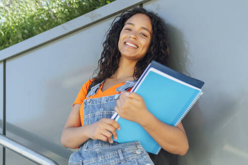 Happy student holding spiral notebooks and leaning on wall in campus - PGF01691