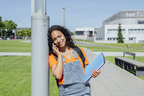 Junge Studentin, die mit ihrem Smartphone spricht und ein Notebook in der Nähe eines Pfostens auf dem Campus hält - PGF01702