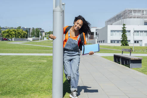 Lächelnder Student, der ein Buch hält und in der Nähe eines Pfostens auf dem Campus steht - PGF01703