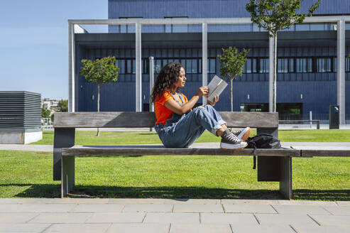 Junge Studentin sitzt auf einer Bank und studiert vor einem Gebäude - PGF01705