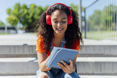 Smiling student wearing wireless headphone and using tablet PC on steps - PGF01708