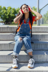 Smiling student listening to music through wireless headphones on steps - PGF01709