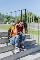 Smiling student with curly hair sitting on steps in campus - PGF01713