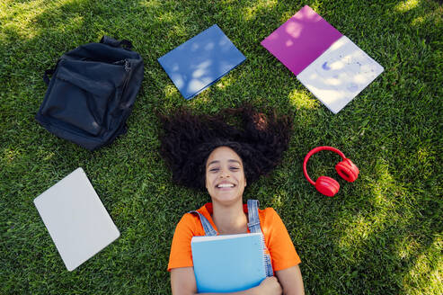 Smiling student lying on grass with spiral notebooks in campus - PGF01717