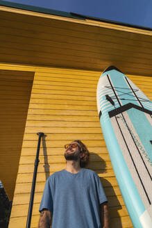 Lächelnder Mann, der in der Nähe eines Paddelboards vor einer Strandhütte steht - VPIF08809