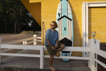 Lächelnder Mann sitzt in der Nähe des Paddelboards vor einer Strandhütte - VPIF08812