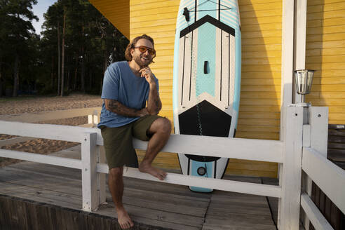 Glücklicher Mann, der in der Nähe eines Paddelboards vor einer Strandhütte sitzt - VPIF08813