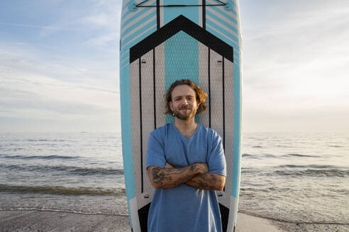 Young man with tattoo standing in front of paddleboard at beach - VPIF08823