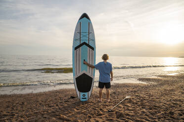 Junger Mann steht in der Nähe Paddleboard auf Sand am Strand - VPIF08832