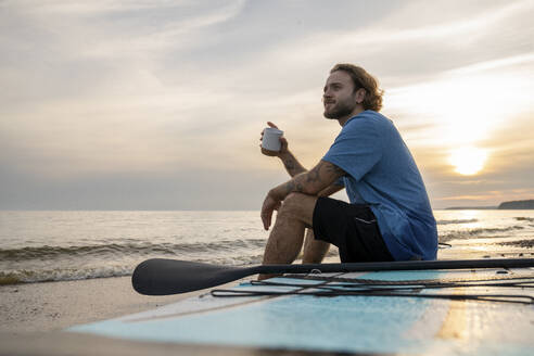 Mann mit Kaffee auf einem Paddelbrett am Strand sitzend - VPIF08843