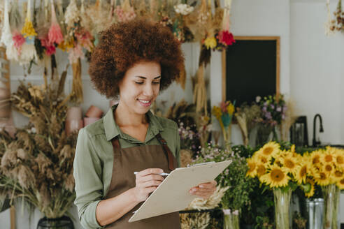 Lächelnde junge Blumenladenbesitzerin beim Schreiben auf einem Klemmbrett im Geschäft - YTF01188
