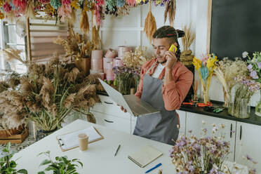 Junger Unternehmer, der in einem Blumenladen mit seinem Smartphone telefoniert und einen Laptop benutzt - YTF01206