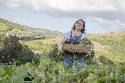 Lächelnde Frau trägt Gemüsekiste unter bewölktem Himmel auf dem Bauernhof - OSF02224