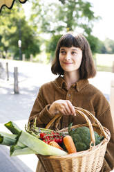 Happy woman wearing brown shirt holding basket of vegetables at farmer's market - AMWF01891