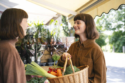 Lächelnde Frau mit Korb voller Gemüse im Gespräch mit ihrer Schwester auf dem Bauernmarkt - AMWF01892