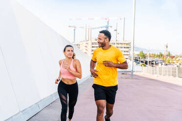 Interracial sportive couple running outdoors - Multiracial couple of runners wearing sportswear and doing functional workout outdoors to strenght body muscles, core abs, stamina and cardio - DMDF07109