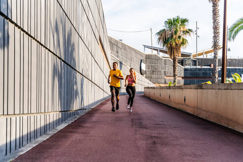 Interrassisches sportliches Paar, das im Freien läuft - Ein gemischtrassiges Läuferpaar trägt Sportkleidung und absolviert ein funktionelles Training im Freien, um die Körpermuskulatur, die Bauchmuskeln, die Ausdauer und das Herz-Kreislauf-System zu stärken - DMDF07116
