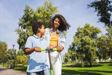 Schöne glückliche afrikanische amerikanische Familie, die sich im Park trifft - Schwarze Familie, die Spaß im Freien hat, Mutter, die ihrem Sohn das Rollerfahren beibringt - DMDF07169