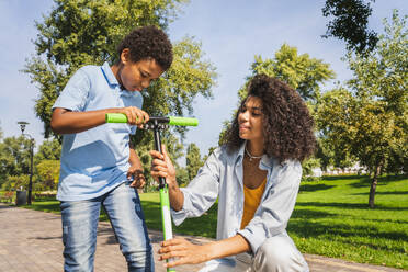 Schöne glückliche afrikanische amerikanische Familie, die sich im Park trifft - Schwarze Familie, die Spaß im Freien hat, Mutter, die ihrem Sohn das Rollerfahren beibringt - DMDF07170