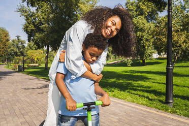 Schöne glückliche afrikanische amerikanische Familie, die sich im Park trifft - Schwarze Familie, die Spaß im Freien hat, Mutter, die ihrem Sohn das Rollerfahren beibringt - DMDF07171