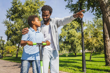 Schöne glückliche afrikanische amerikanische Familie, die sich im Park trifft - Schwarze Familie, die Spaß im Freien hat, Vater, der seinem Sohn das Rollerfahren beibringt - DMDF07172