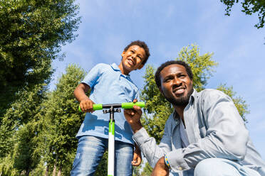 Schöne glückliche afrikanische amerikanische Familie, die sich im Park trifft - Schwarze Familie, die Spaß im Freien hat, Vater, der seinem Sohn das Rollerfahren beibringt - DMDF07173