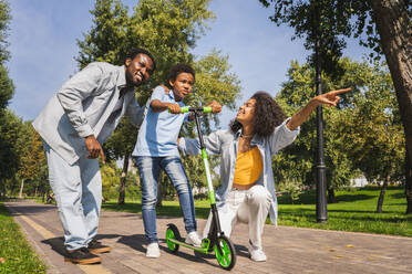 Schöne glückliche afrikanische amerikanische Familie, die sich im Park trifft - Schwarze Familie, die Spaß im Freien hat, Eltern, die ihrem Sohn das Fahren auf dem Roller beibringen - DMDF07175